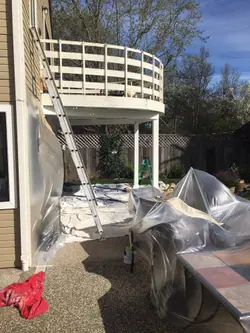 A raised balcony and the patio beneath it masked off before painting, with plastic sheeting taped down the siding, drop cloths across the concrete, garden furniture wrapped, and a ladder set against the balcony edge.