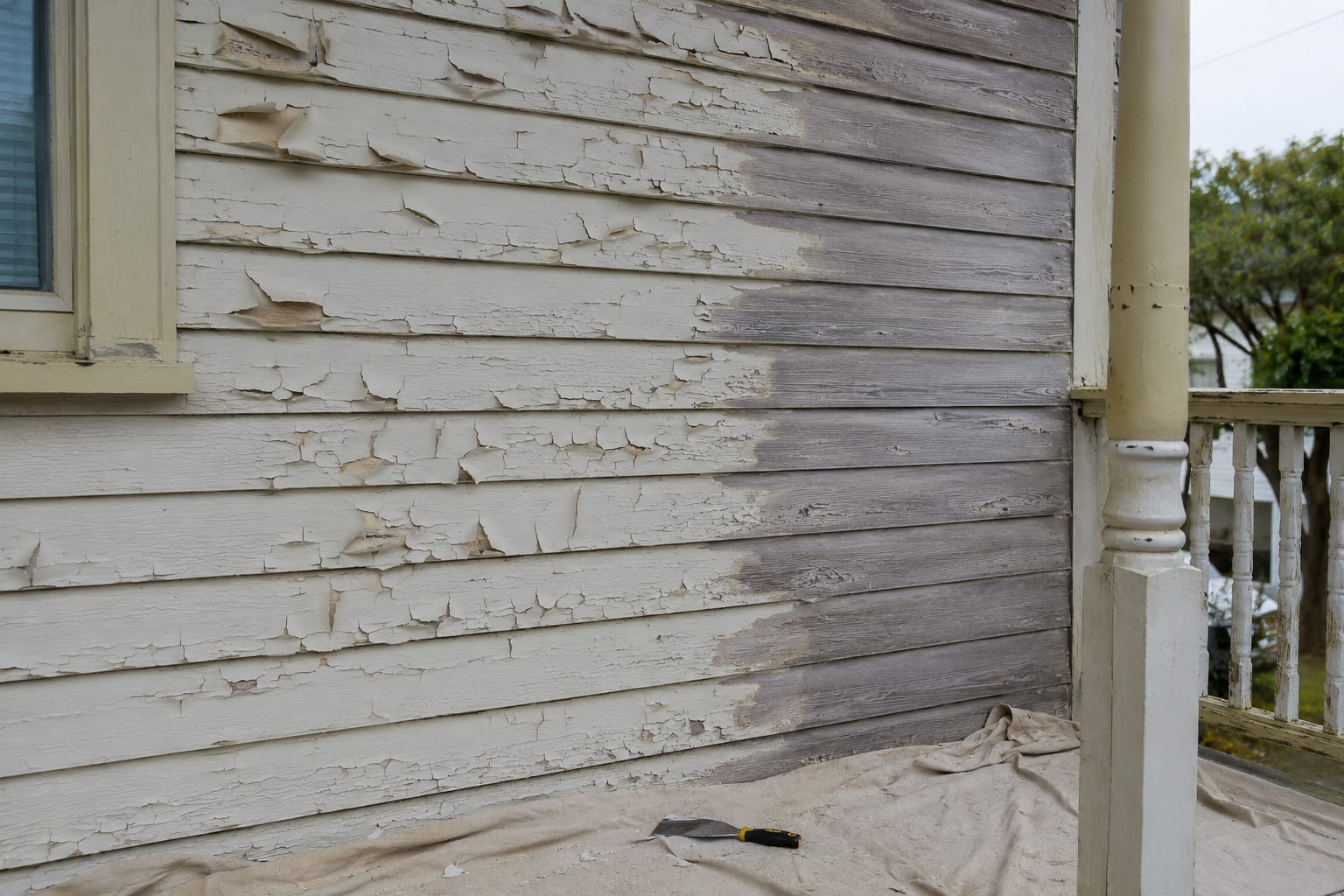 Wooden lap siding with the old white paint lifting and flaking away, part of it already scraped back to bare grey timber, and a dust sheet and scraper on the boards below beside a turned porch railing.