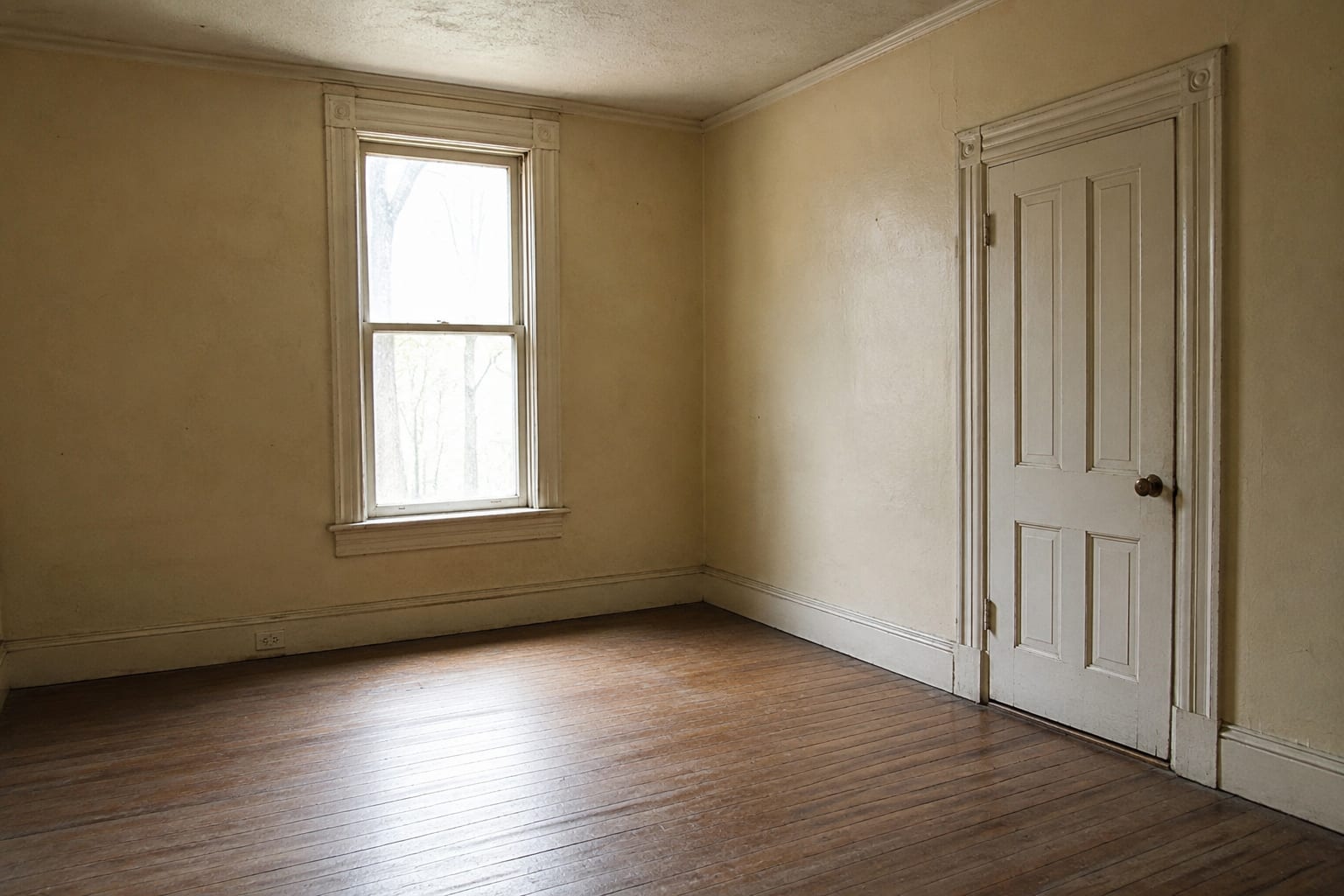 The empty room of an older house, with tall painted window and door casings, deep skirting, aged cream walls and original wood flooring.