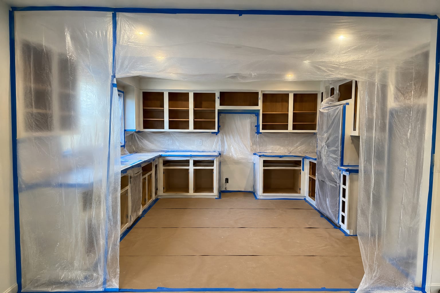 A kitchen sealed off for spraying: clear plastic sheeting taped floor to ceiling across the opening, blue tape along every worktop and cabinet edge, paper covering the whole floor, and the cabinet doors already taken off.