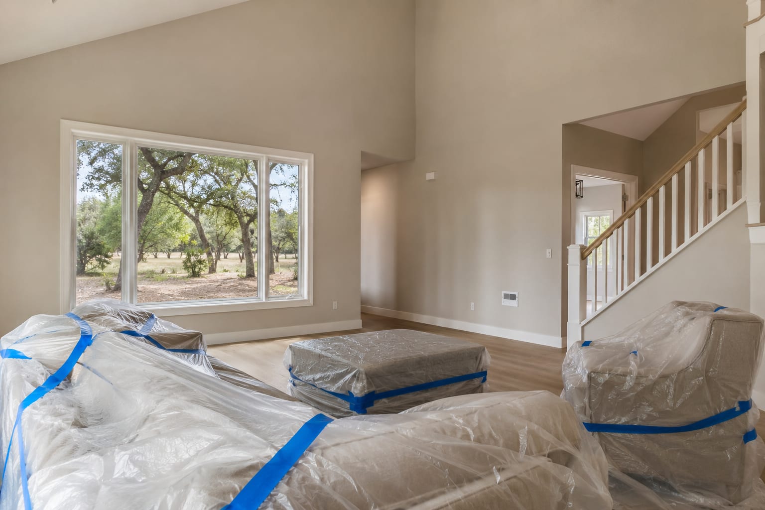 A high-ceilinged living room with the furniture wrapped in plastic sheeting and taped down before painting, a staircase to one side and a wide window looking out onto oak trees.